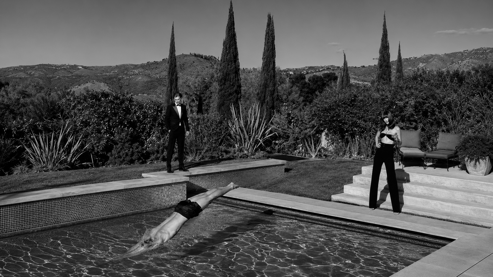 Cypress-lined pool at a hillside estate — a tux at the edge, a woman standing by the steps, a figure caught mid-dive